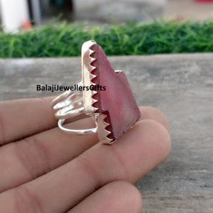May include: A silver ring with a large, irregularly shaped, pinkish-red gemstone. The ring has a triple band design and a serrated edge around the gemstone. The text "BalajiJewellersGifts" is visible on the image.