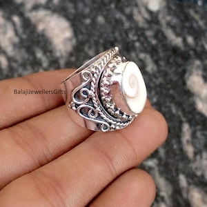 May include: A silver ring with a large, oval, white shell centrepiece. The ring features intricate scrollwork and beaded detailing around the shell. The text "BalajiJewellersGifts" is visible on the side of the ring.