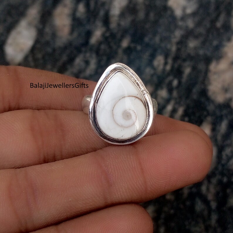 May include: A silver ring with a teardrop-shaped white stone, displaying a spiral pattern. The stone is set in a silver bezel. The ring is shown on a hand. The text "BalajiJewellersGifts" is visible.
