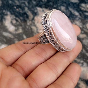 Könnte beinhalten: Ein großer, ovaler Ring mit Rosenquarz-Edelstein und einem verzierten Silber-Filigran-Setting. Der Ring hat einen polierten, hellrosa Stein und aufwendige Details um das Band. Der Text "BalajiJewellersGifts" ist auf dem Ring zu sehen.
