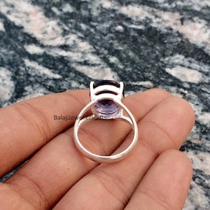 May include: A silver ring featuring a large, oval-cut amethyst gemstone. The amethyst has a deep purple hue at the top, fading to a lighter lavender color. The ring's band is a simple, polished silver design. The text "BalajiJewellersGifts" is visible.