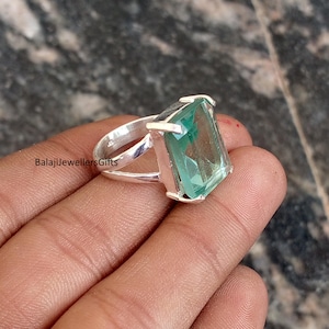 May include: A silver ring with a large, rectangular, light green gemstone. The ring has a split shank design and the text "BalajiJewellersGifts" is visible on the ring's band. The gemstone is set in a silver bezel.