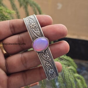 May include: A silver-toned cuff bracelet with intricate floral and scrollwork designs. The bracelet features an oval, light purple gemstone at its center. The text "BalajiJewellersGifts" is visible on the left side of the image. This piece is a statement jewelry item.
