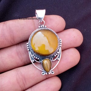 Op de afbeelding: Zilveren hanger met een grote, ronde, goudgele steen en een kleinere, druppelvormige steen eronder. De hanger heeft ingewikkelde zilveren details en een lus voor een ketting. De tekst "BalajiJewellersGifts" is zichtbaar.