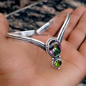 May include: A silver bracelet with a V-shaped design. It features a large, oval-shaped, multi-coloured gemstone and a smaller, round green gemstone. The text "BalajiJewellersGifts" is visible on the bracelet.