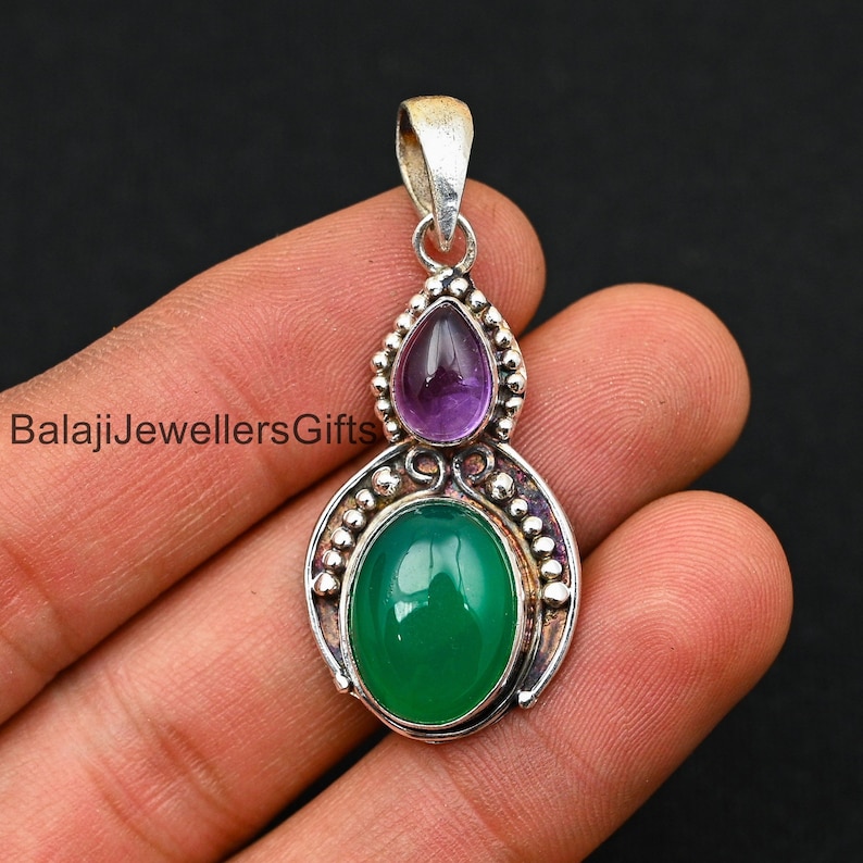 May include: A silver pendant with a green oval gemstone and a smaller purple gemstone. The pendant has a detailed design with small beads around the edges.