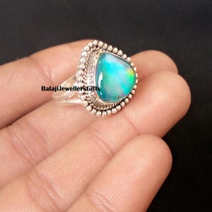 以下が含まれることがあります： 三角形の虹色の宝石が付いたシルバーリング。石は青、緑、黄色の色合いを示しています。リングには、詳細なビーズの縁取りと「BalajiJewellersGifts」の文字があります。