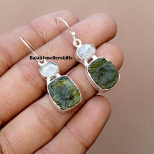 May include: A pair of silver earrings featuring a green, irregularly shaped stone paired with a smaller, light-colored stone. The earrings have a hook closure. The text "BalajiJewellersGifts" is visible.