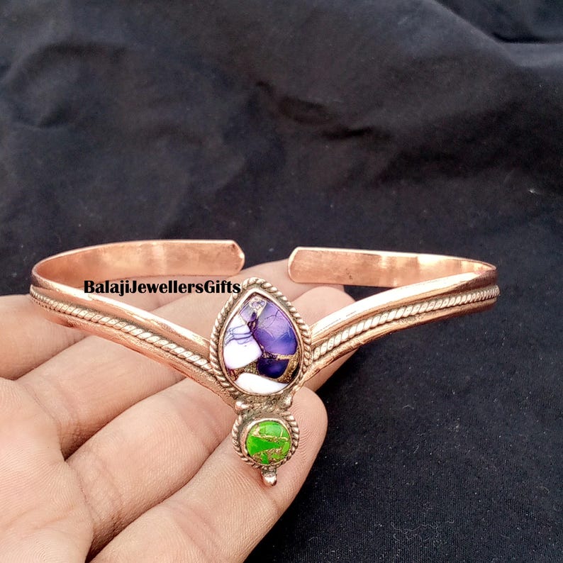 Op de afbeelding: Koperkleurige armband met een centrale druppelvormige steen met paarse, witte en gouden tinten, en een kleinere groene steen eronder. De armband heeft een textuurontwerp en de tekst "BalajiJewellersGifts".