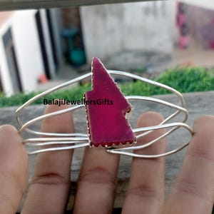 Puede incluir: Una pulsera de brazalete plateada con una vibrante piedra central de color magenta de forma irregular. La piedra está engastada en un marco plateado, y la pulsera tiene un diseño de múltiples hebras. El texto "BalajiJewellersGifts" es visible.