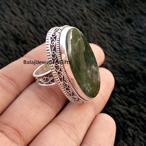 May include: A silver ring with an oval, moss-green stone. The ring has an ornate, filigree design around the stone and on the band. The text "BalajiJewellersGifts" is visible on the ring.