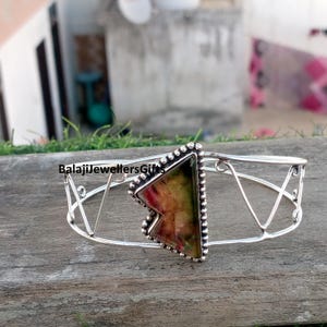 May include: A silver cuff bracelet with a triangular, multi-coloured stone centrepiece. The stone features shades of red, green, and yellow. The bracelet has an open design with geometric cutouts and the text "BalajiJewellersGifts".