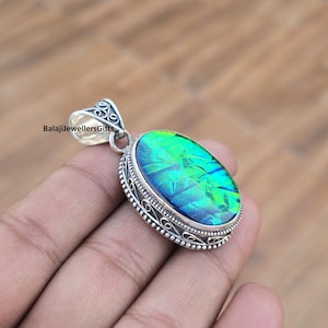 Puede incluir: Colgante ovalado con una piedra preciosa iridiscente en tonos verdes y azules, engastada en un marco de metal plateado con detalles intrincados. El colgante tiene una argolla decorativa para una cadena. El texto "BalajiJewellersGifts" es visible.