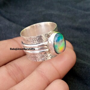 Puede incluir: Un anillo de banda ancha de plata con una piedra cabujón ovalada e iridiscente en tonos azules, verdes y amarillos. La superficie metálica presenta un intrincado patrón floral en relieve y dos bandas horizontales lisas que rodean el centro.