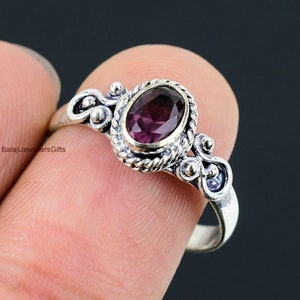 Peut inclure: Une bague en argent avec une pierre précieuse violette sertie dans une bélière dorée. La bague présente un design délicat et orné de volutes et de petites perles.