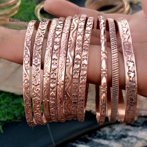 May include: A collection of copper-colored bangles with various embossed designs, including floral, heart, and geometric patterns. The bangles are displayed on a hand, showcasing their intricate details and warm metallic tones. The text "BalajiJewellersGifts" is visible.