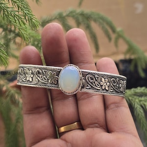 Puede incluir: Un brazalete plateado con una piedra preciosa ovalada de color azul pálido en el centro. La banda del brazalete presenta un diseño floral y de volutas. El texto "Balaji Jewellers & Gifts" es visible en la banda.