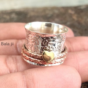 Puede incluir: Un anillo giratorio plateado con un patrón floral en la banda superior y un acento de corazón dorado. El anillo tiene múltiples bandas que giran libremente. El texto "Bala jii" es visible en el lado izquierdo.