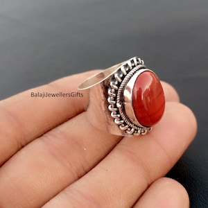 May include: A silver ring with a large, oval, red gemstone. The ring has a decorative border around the stone and the text "BalajiJewellersGifts" is visible. The ring is on a finger.