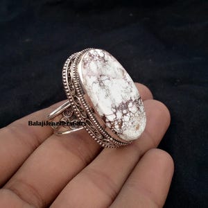Puede incluir: Anillo de plata con una piedra ovalada blanca con vetas marrones. El anillo tiene un diseño de filigrana ornamentado alrededor de la piedra y en la banda. El anillo se muestra sobre un fondo oscuro.
