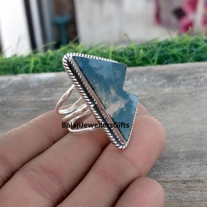 May include: A silver ring with a large, angular, moss agate stone. The stone is a mix of green and white, set in a silver frame with a rope-like border. The ring has a split shank design. The text "BalajiJewellersGifts" is visible.
