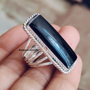 May include: A silver ring with a large, rectangular black stone. The ring has a braided border around the stone and multiple bands on the ring's base. The text "BalajiJewellersGifts" is visible on the ring.