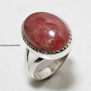 Pode incluir: Um anel de prata com uma grande pedra preciosa ovalada, rosa avermelhada. A pedra é definida em uma moldura texturizada. A banda do anel divide-se em duas partes. O texto "BalajiJewellersGifts" é visível.
