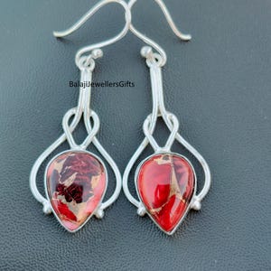 May include: A pair of silver-toned dangle earrings. Each earring features a teardrop-shaped, red and gold-toned stone set in a silver frame. The earrings have a unique, looping design above the stone and a hook closure. The text "BalajiJewellersGifts" is visible.