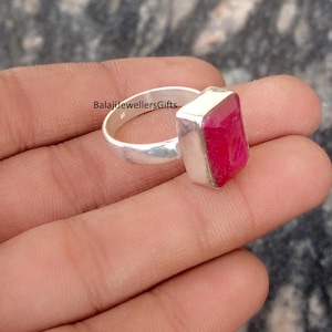 Puede incluir: Un anillo de plata con una piedra preciosa roja vibrante de forma rectangular. La banda del anillo es lisa y pulida, y la piedra preciosa está engastada en un bisel de plata. El texto "BalajiJewellersGifts" está grabado en la banda del anillo. Este anillo es una joya.