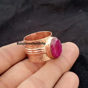 May include: A wide copper ring featuring a textured, embossed surface pattern and a prominent, oval-shaped, faceted deep pink gemstone set in a smooth bezel. The text BalajiJewellersGifts is visible on the band of this metallic ring.
