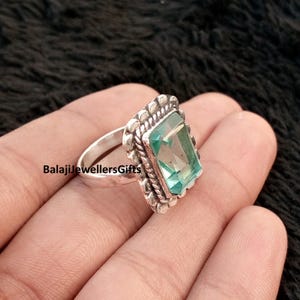 May include: A silver ring with a rectangular, light blue gemstone. The gemstone is set in a decorative, textured frame. The ring is on a hand, with the text "BalajiJewellersGifts" visible.