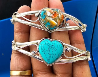 May include: Two silver heart-shaped bracelets with turquoise stones. The top bracelet has a brown and orange stone, while the bottom bracelet has a blue stone.