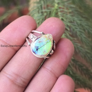 Op de afbeelding: Een zilveren ring met een peervormige, iriserende edelsteen. De edelsteen vertoont een reeks kleuren, waaronder groen, blauw en geel. De ring heeft een gedetailleerde band met decoratieve elementen. De tekst "BalajiJewellersGifts" is zichtbaar.