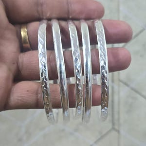 Może przedstawiać: Zestaw srebrnych bransoletek o młotkowanej fakturze. Bransoletki są prezentowane w dłoni, podkreślając ich odblaskową powierzchnię i skomplikowane detale. Bransoletki są w kolorze jasnego srebra.