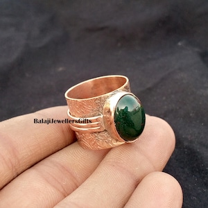 May include: A copper-colored ring with an oval, dark green gemstone. The ring has an engraved floral pattern and the text "BalajiJewellersGifts". The ring is a wide band style.