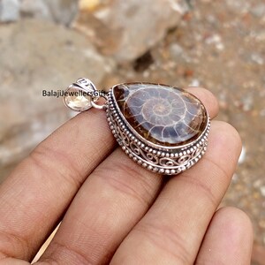 May include: A teardrop-shaped pendant with a polished, brown fossil set in a silver filigree frame. The fossil displays a spiral pattern. The pendant has a small loop at the top for a chain. The text "BalajiJewellersGifts" is visible.