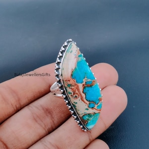 May include: A silver ring with a large, elongated turquoise stone. The stone features a mix of blue, brown, and cream colors. The ring's band is silver, and the stone is surrounded by a beaded silver border. The text "BalajiJewellersGifts" is visible.