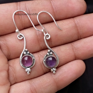 Puede incluir: Un par de pendientes plateados con un diseño de lágrima. Cada pendiente presenta una piedra preciosa redonda, de color púrpura oscuro, engastada en un bisel decorativo. Los pendientes tienen un gancho curvado para orejas perforadas.