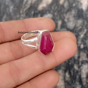 May include: A silver ring with a large, faceted, ruby-red gemstone. The ring has a double band design and the text "BalajiJewellersGifts" is visible. The gemstone is a vibrant, eye-catching focal point.