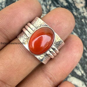 以下が含まれることがあります： 長方形のフレームにセットされた、楕円形の赤みがかったオレンジ色の宝石が付いたシルバーリング。 リングには装飾的な花の模様と、宝石の両側に3つの水平バンドがあります。