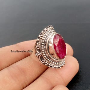 May include: A silver ring with an oval, faceted ruby gemstone. The ring has an ornate, textured setting with a beaded border and scrollwork details. The text "BalajiJewellersGifts" is visible.
