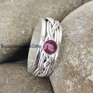 May include: A silver ring with a braided design and a red gemstone in the center. The ring is set against a background of gray rocks.