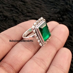 May include: A silver ring with a large, emerald-cut green gemstone. The ring has a detailed, textured setting with a rope-like border and small bead accents. The ring is displayed on a dark background. The text "BalajiJewellersGifts" is visible.