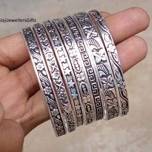 Könnte beinhalten: Ein Set aus sieben silberfarbenen Armreifen mit aufwendigen floralen und geometrischen Mustern. Die Armreifen werden in einer Hand gehalten, um die detaillierten Designs zu zeigen. Die Armbänder haben eine Vintage- oder Antik-Ästhetik.