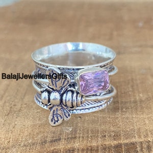 May include: A silver spinner ring with a rectangular, pale pink gemstone. The ring features a detailed bee design and the text "BalajiJewellersGifts". The ring has multiple bands that spin freely.