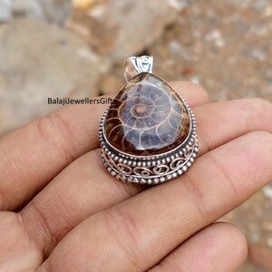May include: A silver pendant with a teardrop-shaped, polished brown fossil. The fossil features a spiral pattern and is set in a decorative silver bezel. The text "BalajiJewellersGift" is visible on the left side of the pendant.