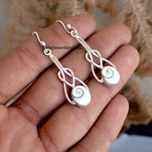 Peut inclure: Boucles d'oreilles en argent au design unique. Chaque boucle d'oreille présente une longue tige argentée avec un détail en forme de nœud et une pierre ovale blanche avec un motif en spirale. Le texte "BalajiJewellersGifts" est visible.