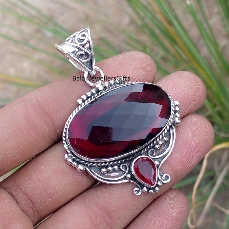 May include: A silver pendant with a large, faceted, oval-shaped, deep red gemstone as the focal point. A smaller, teardrop-shaped red gemstone is below. The pendant has ornate silver detailing and the text "BalajiJewellersGifts".