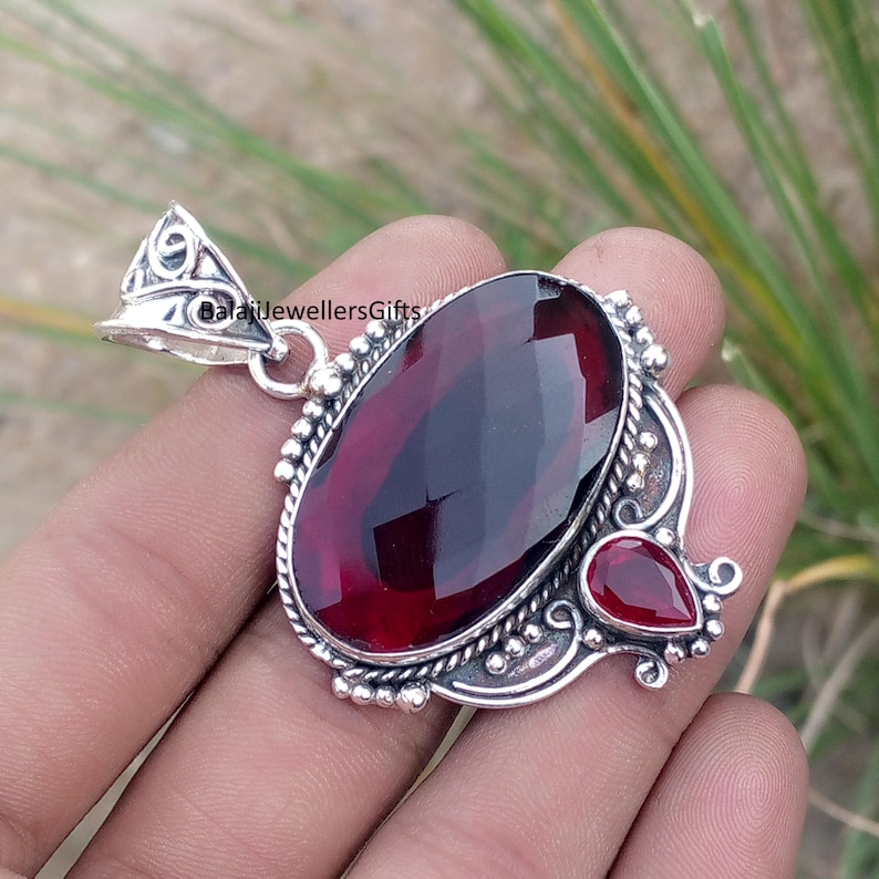 May include: A silver pendant with a large, oval-cut, deep red gemstone as the focal point. A smaller, teardrop-shaped red stone is set to the side. The pendant has an ornate, detailed silver setting and a bail for a chain.