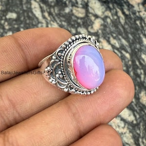 Puede incluir: Un anillo de plata con una piedra preciosa ovalada e iridiscente. La piedra preciosa cambia de rosa a lavanda. El anillo tiene un engaste ornamentado y detallado con un borde de cuentas. El texto "BalajiJewellersGifts" es visible.